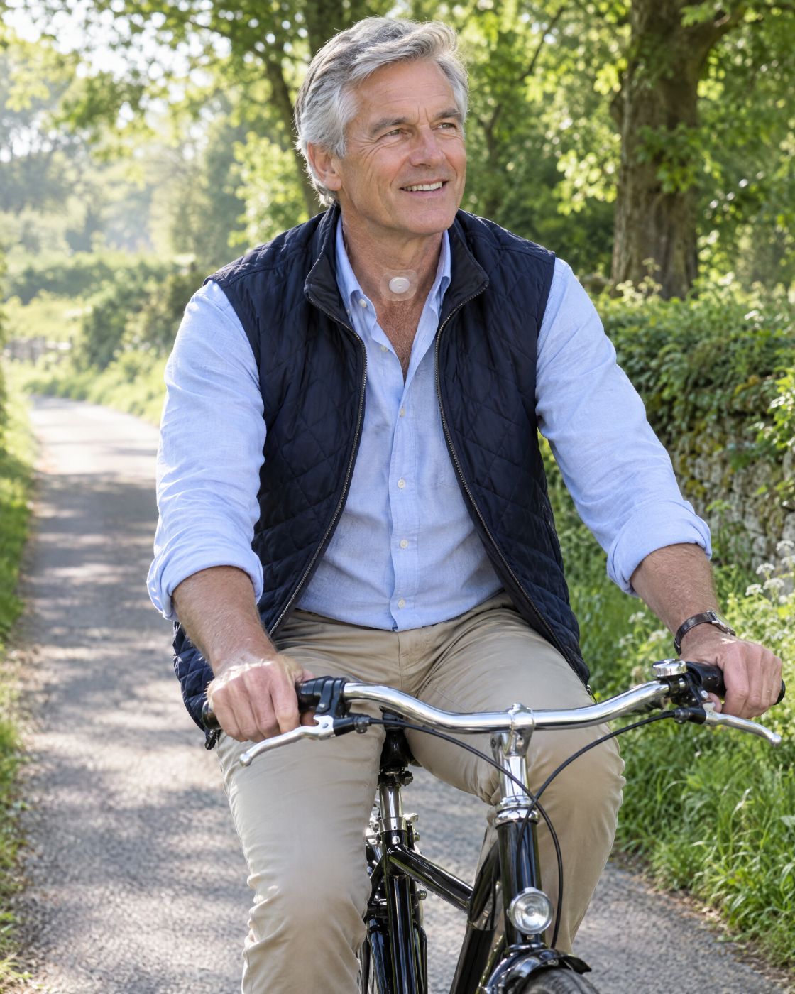 A grey-haired man in a navy gilet and blue shirt rides a bicycle along a tree-lined lane, an HME visible at the base of his neck.