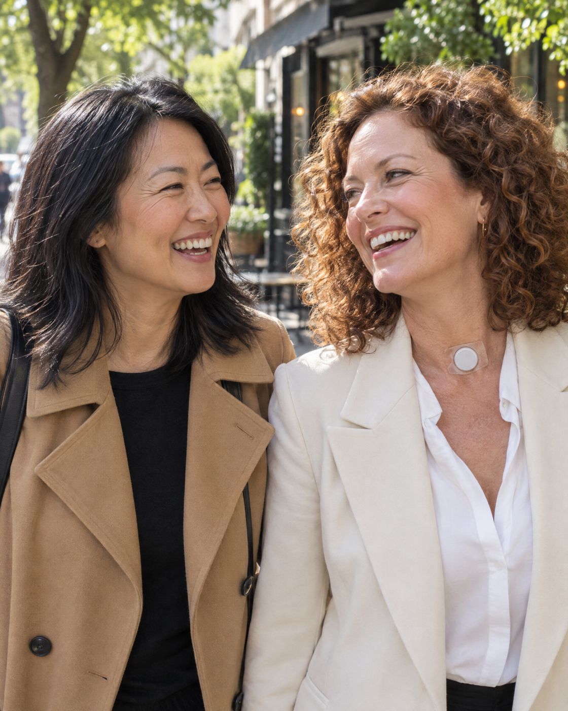 Two friends in their forties laugh together on a sunny street. One wears an HME at the base of her neck.